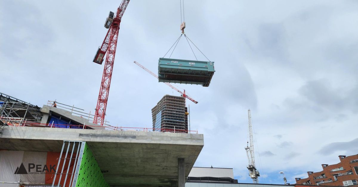 Crane lifting a green construction dumpster high above a busy urban building site in BC.