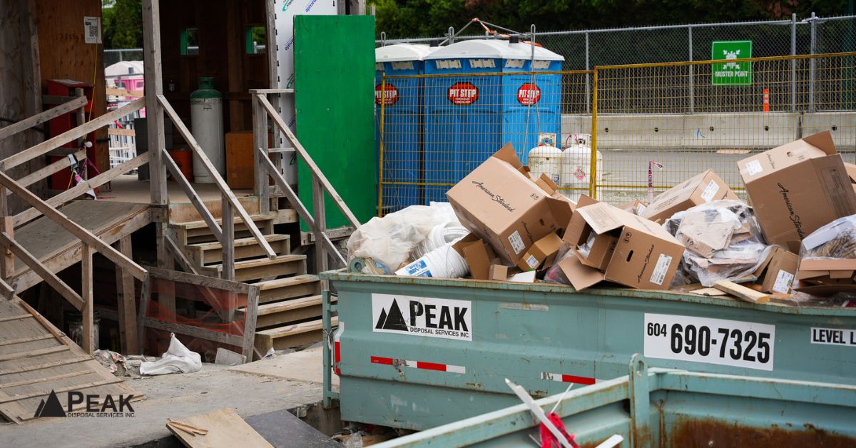 Peak Disposal bin filled with cardboard and construction debris sits next to wooden stairs and portable toilets at a worksite