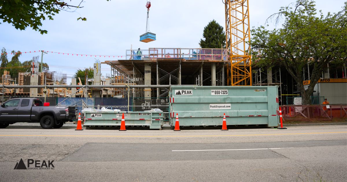 green Peak Disposal bin placed on a street beside a busy construction site with crane and truck.