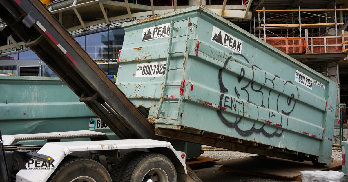 Large green roll-off dumpster being lifted and tilted by a white disposal truck at an active construction site.