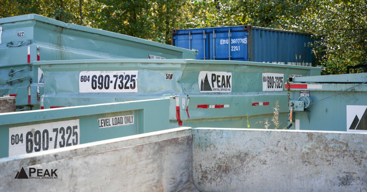 Several light green Peak Disposal roll-off bins arranged in a storage yard with trees and containers.
