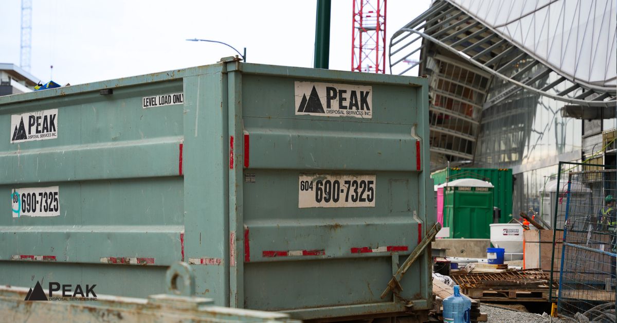 Peak Disposal Services bin marked 'LEVEL LOAD ONLY' positioned beside modern buildings and construction materials in Abbotsford