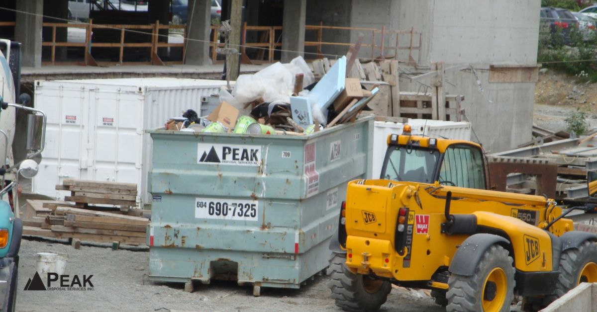 teal Peak Disposal bin next to a yellow JCB telehandler at a crowded urban construction site in BC.