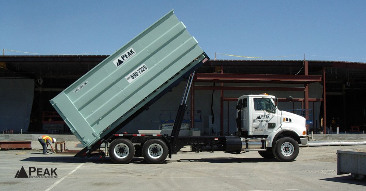 White Peak Disposal roll-off truck tilting a large teal bin to dump heavy materials at an industrial yard.