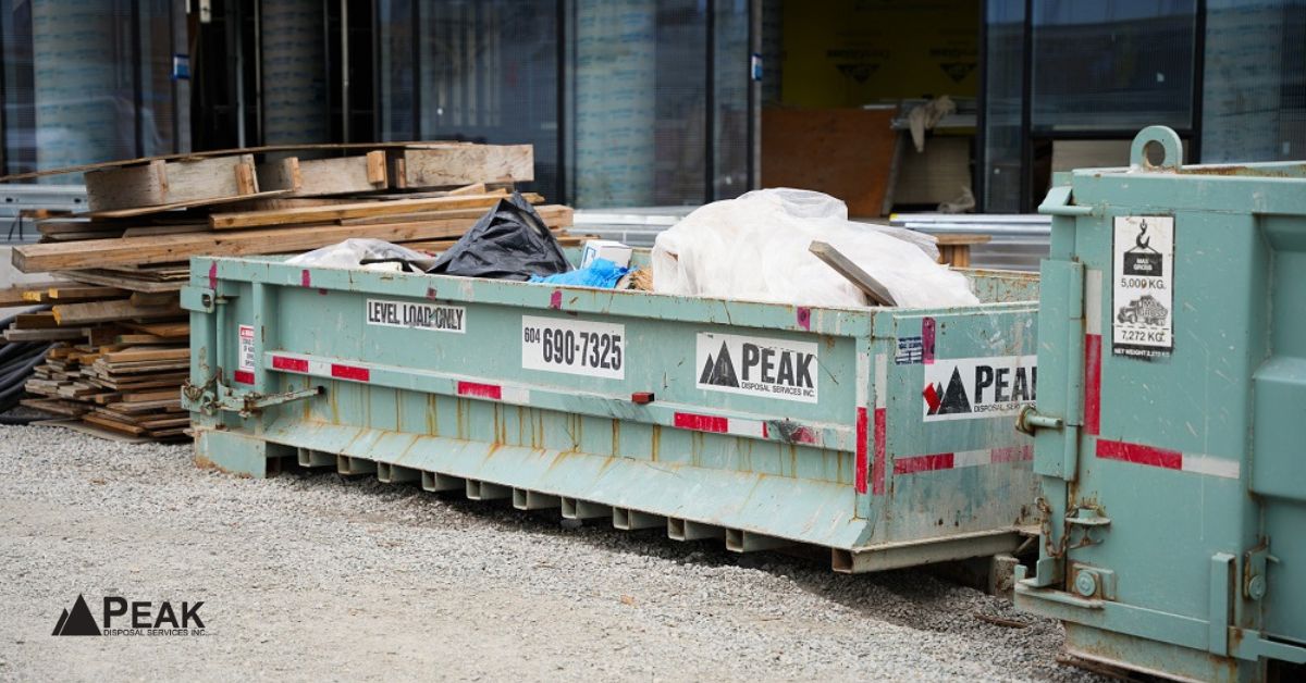 Large green Peak construction dumpster filled with wood scraps and debris on a gravel site next to another bin