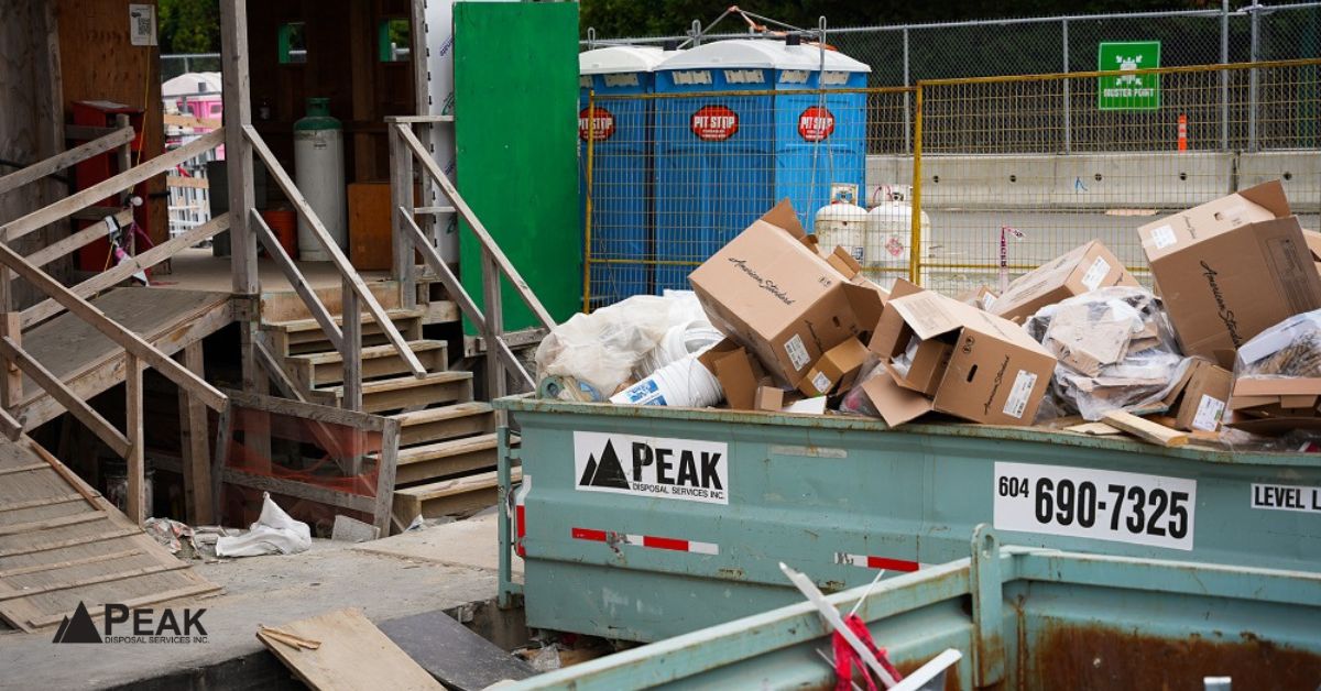 Overflowing green Peak dumpster filled with cardboard boxes and debris at a busy construction site with wooden stairs