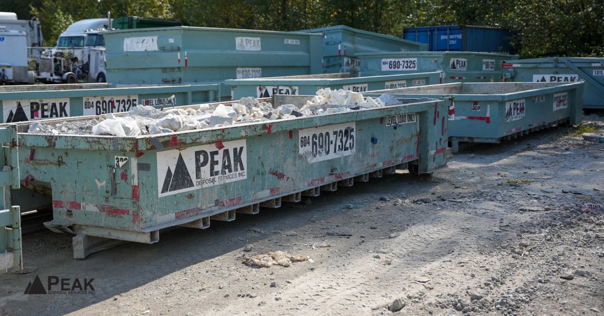 Multiple large green Peak Disposal dumpsters filled with construction debris and concrete waste lined up in a gravel yard
