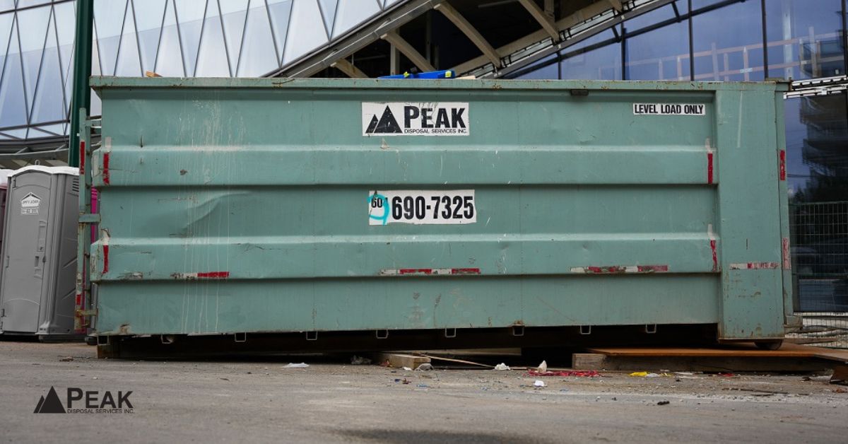 Peak Disposal dumpster positioned on pavement in front of a modern glass building, with portable toilet nearby