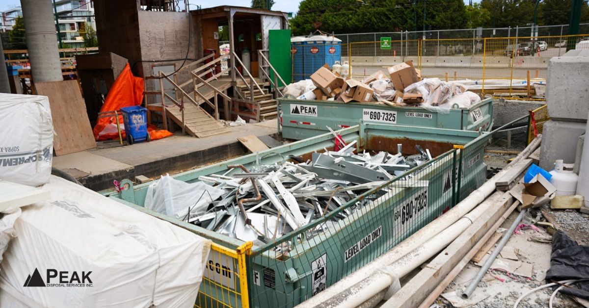 Busy construction site with large green Peak dumpsters overflowing with scrap metal, cardboard, and mixed debris amid active building work