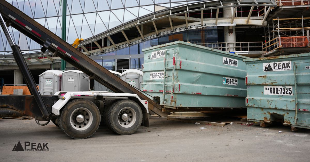Peak Disposal roll-off truck delivering large green dumpsters at a Metro Vancouver construction site with modern building in background.