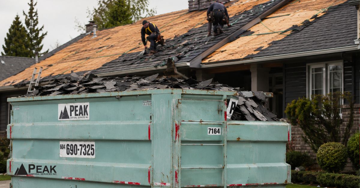 Roofing dumpster filled with asphalt shingles beside a residential roof replacement project with workers above.