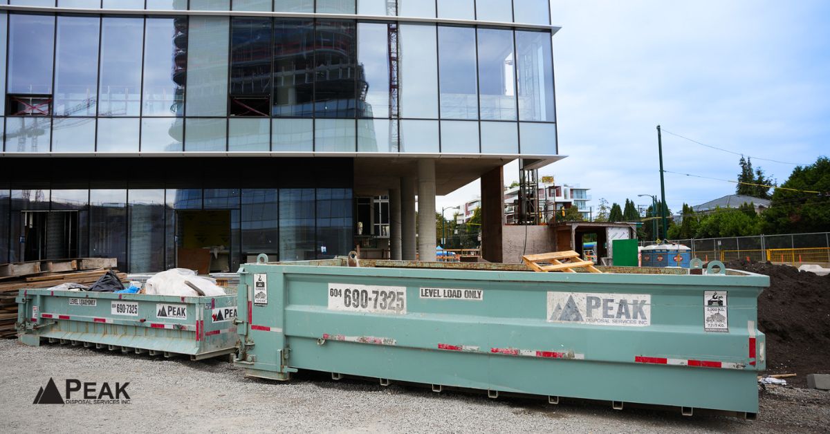 Green PEAK disposal dumpsters at an active construction site with a modern glass building in the background