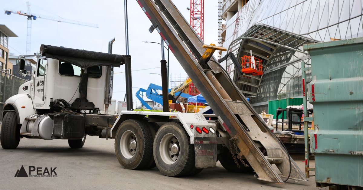 PEAK Disposal white roll-off truck hoisting a green dumpster at a busy urban construction site with cranes and modern buildings