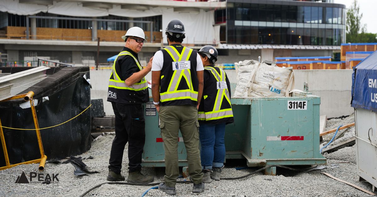 Contractors review hauling plans beside an empty roll-off bin, excavator, soil piles, and concrete rubble onsite.