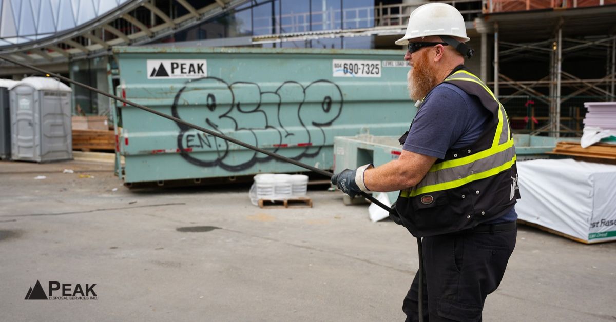 Peak Disposal worker coordinates heavy-material hauling beside a construction dumpster at an active jobsite.