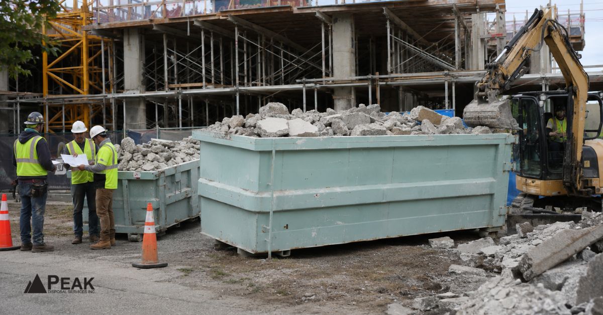 Workers review concrete removal plans beside two loaded bins, excavator, rubble, and active construction site operations.